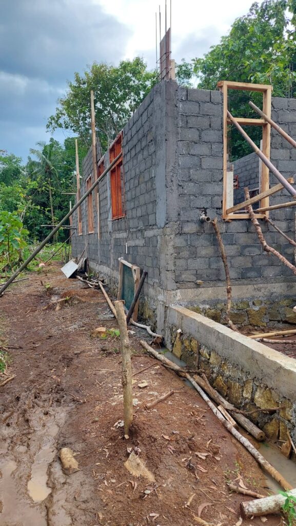 Rohbau eines Hauses ohne Dach und mit unverputzten Wänden und Fensterrahmen aus Holz, in die noch keine Fenster eingesetzt sind und die mit langen Bambusstangen gestützt werden. Der Boden ringsrum ist matschig.