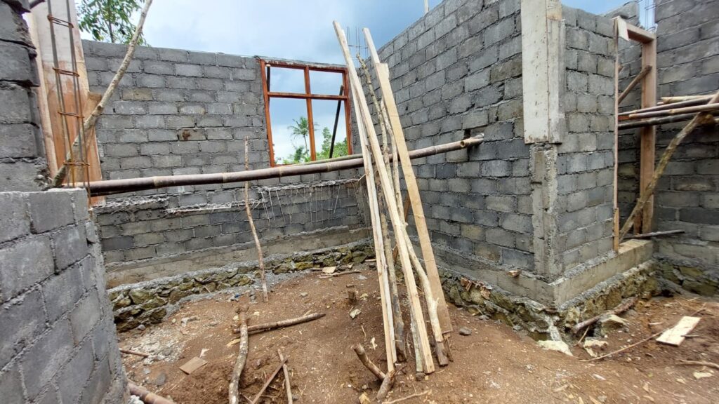 Ein halbfertiges Zimmer ohne Dach mit unverputzten Wänden aus grauen Mauersteinen, an den Ecken ragt Baustahl heraus, die Fenster- und Türrahmen aus Holz sind leer, es liegen Holzlatten und Bambusstangen herum, der Boden besteht aus unebener festgetretener Erde.