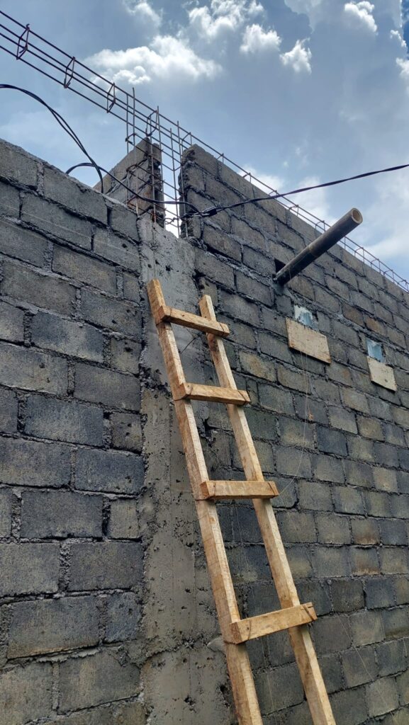 An einer unverputzten Wand aus grauen Mauersteinen lehnt eine aus unbearbeiteten Holzlatten gezimmerte Leiter, zur Mauer führt ein Stromkabel, darauf liegt eine Art längliche viereckige Röhre aus Metallstangen.